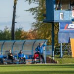 Zadaszone siedziska na stadionie sportowym. Na czterech siedzą mężczyźni w sportowych strojach. Obok wieża z wynikiem meczu