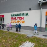 Grupa dzieci w kurtkach i czapkach idze wzdłuż budynku z napisem "Akademia małych zuchów"