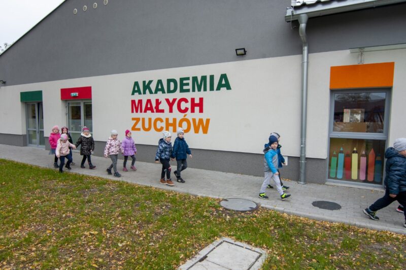 Grupa dzieci w kurtkach i czapkach idze wzdłuż budynku z napisem "Akademia małych zuchów"