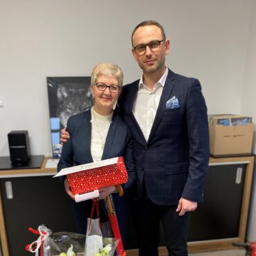 Mężczyzna w granatowej marynarce i okularach obejmuje kobietę w okularach trzymającą kolorowy karton